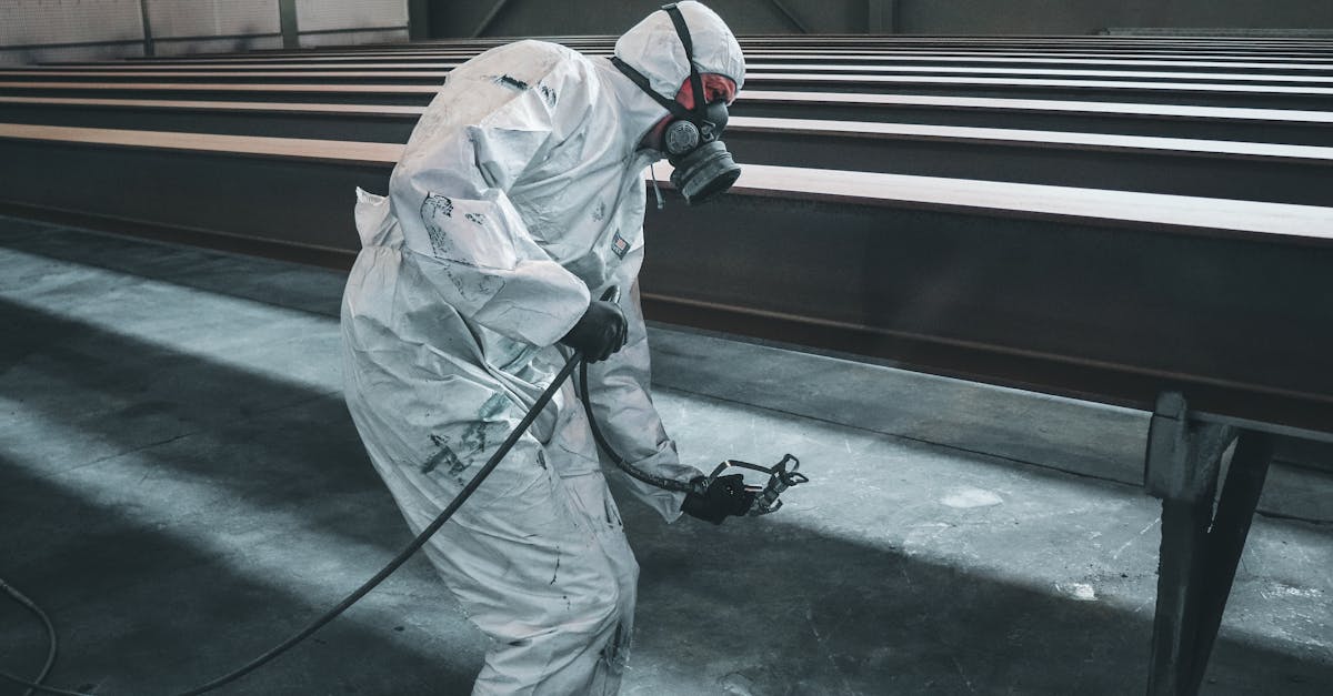 Worker in protective gear spray painting metal beams inside a factory setting.